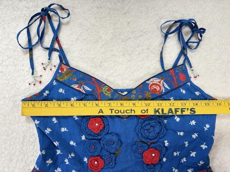 May include: Blue floral print top with red and blue crochet flower accents. The top has tie straps and a decorative floral trim. The text "A Touch of KLAFF'S" is visible on a yellow ruler.
