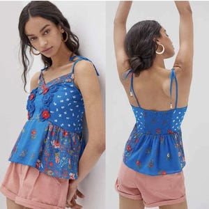 May include: A blue floral print top with a V-neckline and adjustable tie straps. The top features a peplum hem and is adorned with red and blue floral appliqu&eacute;s. The fabric has a white floral pattern and a shirred back panel.