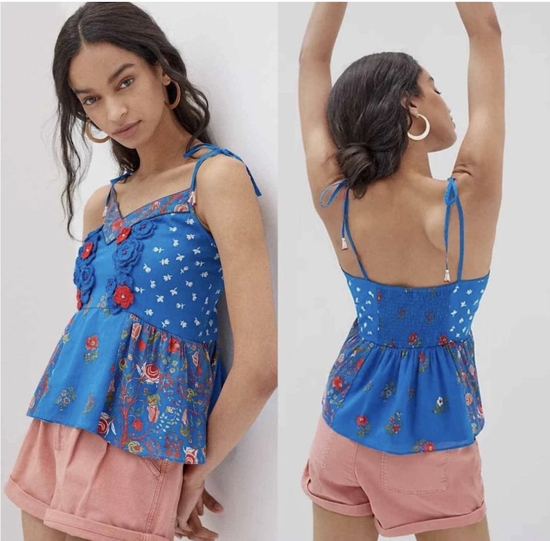 May include: A blue floral print top with a V-neckline and adjustable tie straps. The top features a peplum hem and is adorned with red and blue floral appliqu&eacute;s. The fabric has a white floral pattern and a shirred back panel.