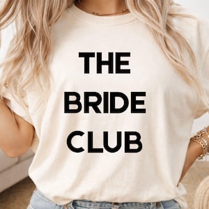 May include: A cream-coloured t-shirt featuring the text "THE BRIDE CLUB" in bold, black capital letters. The shirt is made of a soft material, suitable for casual wear. The design is centered on the front of the shirt.