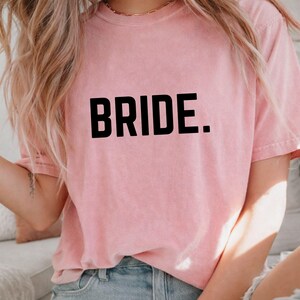May include: A pale pink t-shirt featuring the word "BRIDE." in large, black, capital letters. The shirt has a casual style with short sleeves. The material looks soft and has a slightly worn appearance.