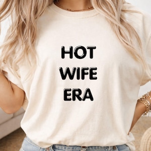 May include: A cream-coloured t-shirt featuring the text "HOT WIFE ERA" in bold, black lettering. The shirt is made of a soft material and has a crew neck. The image shows the upper body of a person wearing the t-shirt.