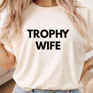 May include: A cream-coloured t-shirt featuring the text "TROPHY WIFE" in bold, black capital letters. The shirt has a crew neck and short sleeves, with a relaxed, casual style.