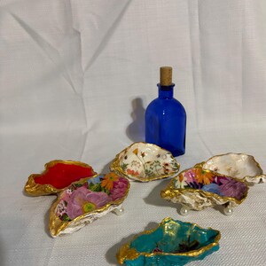 May include: A collection of decorative oyster shell dishes with gold trim and floral designs. A cobalt blue glass bottle with a cork stopper is also present. The shells are painted in various colors, including red, teal, and white.