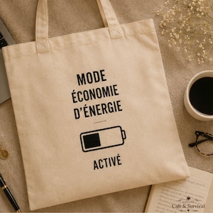 Peut inclure: Un sac fourre-tout en toile beige avec le texte noir MODE ÉCONOMIE D'ÉNERGIE ACTIVÉ imprimé au centre, accompagné d'une icône de batterie partiellement remplie. Le sac est doté de deux anses et est fabriqué dans un tissu de couleur claire.
