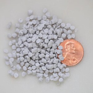 May include: A pile of small, light gray plastic pellets next to a copper penny. The pellets are cylindrical and scattered, with the penny providing a size reference. The penny shows the profile of a man.