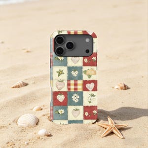 Peut inclure: Coque de téléphone avec un motif patchwork de fraises, de cœurs et de motifs floraux rouges, bleus et crème. L'étui a une découpe pour l'appareil photo et est présenté sur une plage de sable avec des coquillages et une étoile de mer.