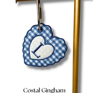 Op de afbeelding: Een blauw en wit geruit hartvormig sleutelhanger met een wit hart in het midden, met de letter "I". De sleutelhanger is bevestigd aan een goudkleurige metalen clip en is voorzien van het label "Costal Gingham".