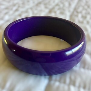 May include: A glossy, solid purple bangle bracelet. The wide, circular bracelet has a smooth, polished surface. The interior of the bracelet is a darker shade of purple. The bracelet is a fashion accessory.
