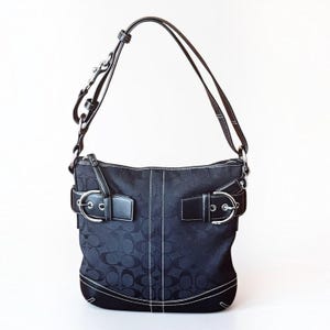 May include: A black shoulder bag featuring a repeating logo pattern on the fabric body, accented with dark leather trim and silver-tone metal buckles on the front. The bag includes an adjustable shoulder strap and white contrast stitching throughout.