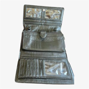 May include: Black leather wallet, open to display multiple compartments. Features include clear ID windows, card slots, and a zippered pocket. The wallet has a snap closure and an embossed design on the front.