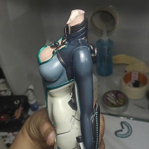 K&ouml;nnte beinhalten: Ein kopfloser Actionfiguren-Torso in einem dunkelblauen und wei&szlig;en Anzug mit t&uuml;rkisfarbenen Akzenten. Die Figur hat einen hohen Kragen, ein eng anliegendes Oberteil und lange &Auml;rmel mit schwarzen Unterarmsch&uuml;tzern sowie detaillierte Linien.