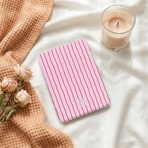 Può includere: Una custodia rettangolare per e-reader con sfondo rosa chiaro e sottili strisce verticali rosa scuro. Il nome Ellie è stampato in un semplice carattere corsivo nero vicino al centro inferiore della custodia dagli angoli arrotondati.