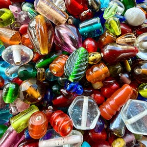 Op de afbeelding: Een grote verzameling diverse glaskralen in verschillende vormen, maten en kleuren, waaronder rood, blauw, groen, oranje, paars en transparant. De kralen hebben diverse texturen, gefacetteerde oppervlakken en gladde afwerkingen met zichtbare gaten.