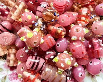 Puede incluir: Una colección de cuentas de vidrio rosas y color rosa en varias formas y tamaños. Algunas cuentas tienen rayas, puntos u otros elementos decorativos. Las cuentas se muestran en un plato con estampado floral.