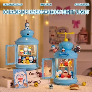 May include: Two light blue Doraemon-themed night lights with openable doors, showcasing miniature figures and food items. The lamps have a lantern design with a top handle and a small Doraemon figure on top. The text "DORAEMON HANDMADE DIY NIGHT LIGHT" is visible.