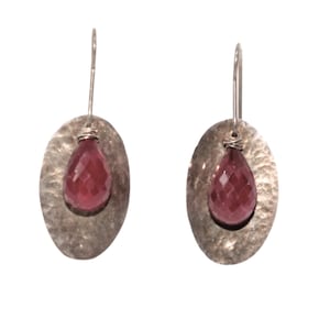 May include: A pair of silver earrings. Each earring features a hammered oval-shaped silver base with a faceted, teardrop-shaped, deep red gemstone suspended in the center. The earrings have a simple hook closure.