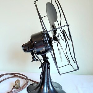 May include: An antique black electric fan with a protective wire cage. The fan has a rounded base and a tilting head. The power cord is brown. The fan blades are visible inside the cage. A vintage home decor item.