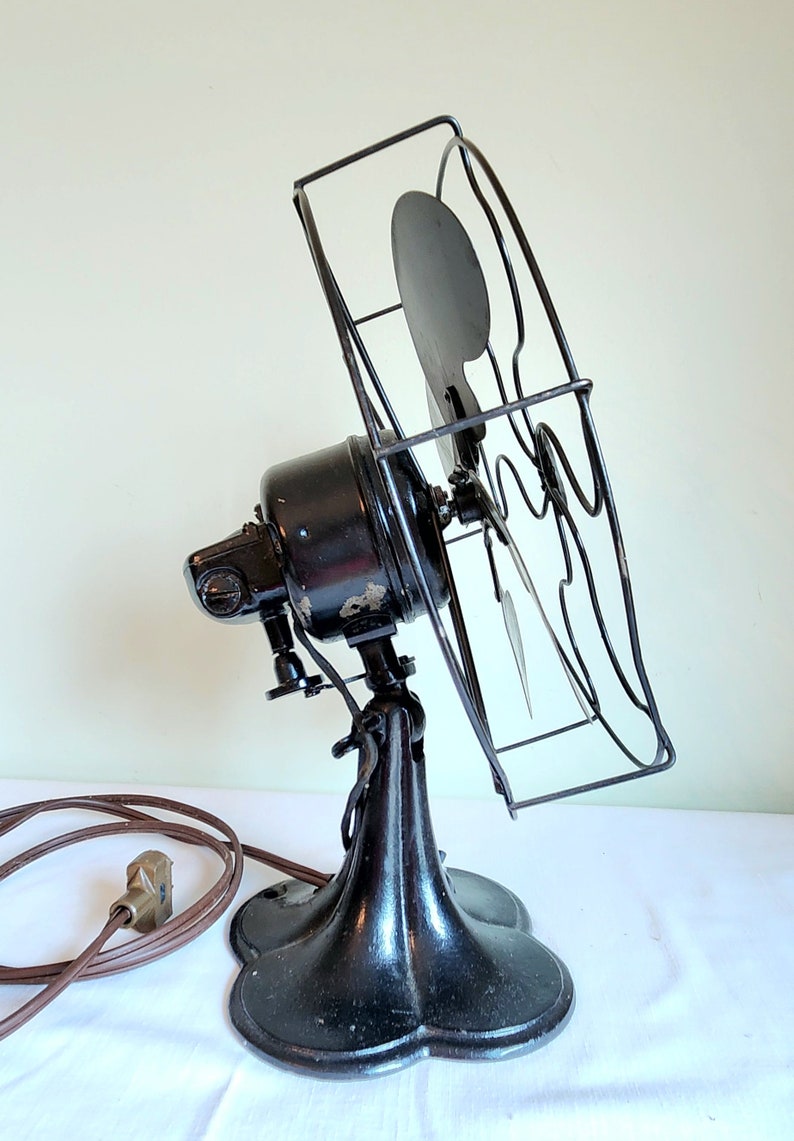 May include: An antique black electric fan with a protective wire cage. The fan has a rounded base and a tilting head. The power cord is brown. The fan blades are visible inside the cage. A vintage home decor item.