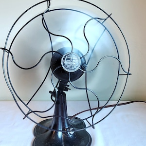 May include: An antique Emerson Seashore electric fan. The fan has a black metal base and cage, with a silver emblem in the center. The fan blades are visible behind the cage. The fan is on a white surface.