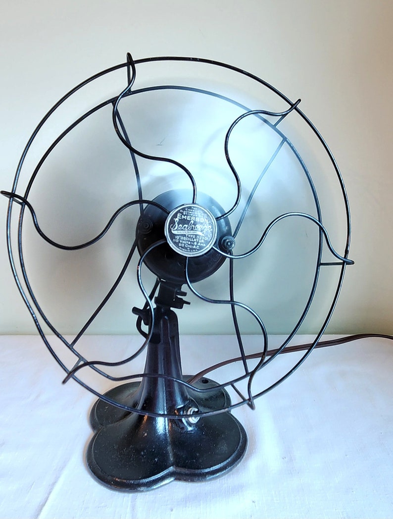 May include: An antique Emerson Seashore electric fan. The fan has a black metal base and cage, with a silver emblem in the center. The fan blades are visible behind the cage. The fan is on a white surface.