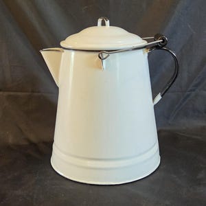 May include: A white enamel coffee pot with a tapered body, a matching lid, and a black handle. The pot features a black rim around the top and base, a side spout, and a metal wire bail handle attached to the sides for carrying or hanging.