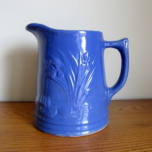 May include: A cobalt blue ceramic pitcher with a handle and a spout. The pitcher features embossed floral designs on the front. The pitcher is sitting on a wooden surface.