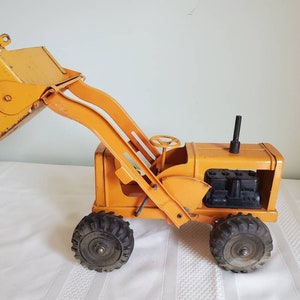 May include: A vintage yellow toy backhoe with a black engine and black tires. The backhoe has a raised arm and a large bucket.