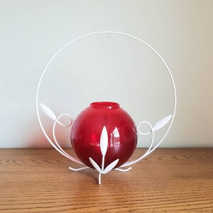 May include: A white metal vase with a red glass globe inside. The vase has a circular design with leaf-like accents.