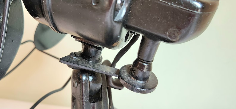 May include: Close-up of a vintage black electric fan. The fan's motor housing and stand are made of dark metal. The image highlights the fan's mechanical details, including the motor housing, blades, and the stand's adjustable features.