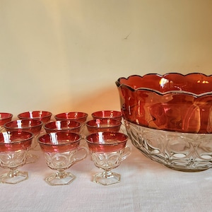 May include: A vintage punch bowl set with a clear glass bowl with a red rim and 12 matching cups. The bowl has a scalloped edge and a textured design. The cups have a red rim and a clear base.