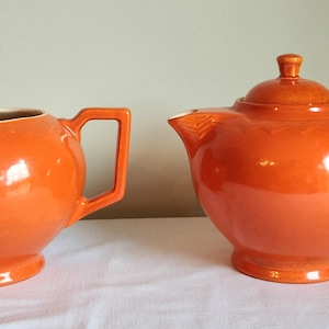 May include: A set of orange ceramic kitchenware, including a teapot and creamer. The teapot has a lid and a handle. The creamer has a handle.