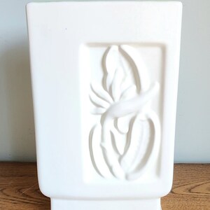 May include: White ceramic vase with a raised design of a stylized flower. The vase has a square body and a rectangular base.