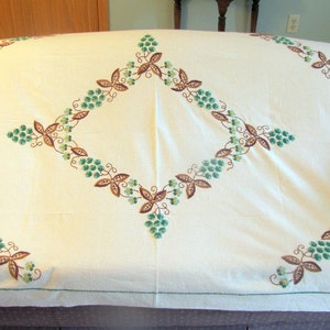 May include: Cream-colored tablecloth with embroidered floral and leaf patterns in shades of brown and green. The design is arranged in a diamond shape in the center and along the edges. The tablecloth is draped over a sofa.