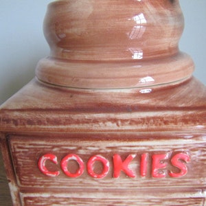 May include: A brown ceramic cookie jar shaped like a wooden drawer with the word "COOKIES" in red letters on the front.