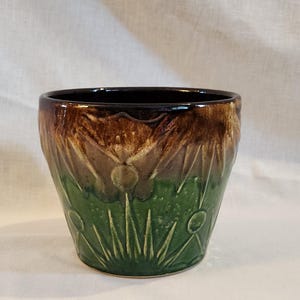 May include: A ceramic planter with a glossy black rim, transitioning to brown and green tones. The planter features a carved floral design, adding a decorative touch. Ideal for indoor plants, this piece is a blend of functionality and art.