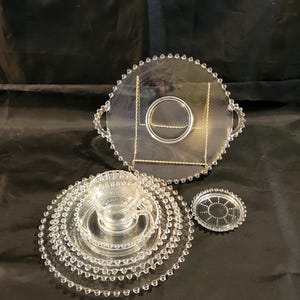 May include: A clear glass serving set with beaded edges. The set includes a tiered serving tray with a gold-colored metal stand, a teacup, saucers, and a coaster. The pieces are arranged on a dark surface.
