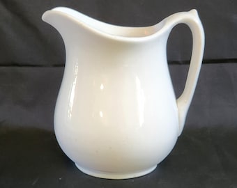 May include: A small, white ceramic creamer pitcher with a curved spout and a rounded body. The handle is a simple loop. The creamer is on a dark background.