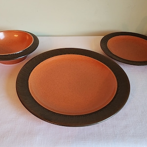 May include: Three ceramic dishes in a set. The dishes are orange with a brown rim. The dishes are on a white tablecloth.