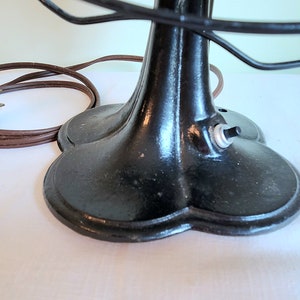 May include: Close-up of a vintage black electric fan. The fan has a round base with a three-leaf design, a black cage, and a brown power cord. The on/off switch is visible on the base.