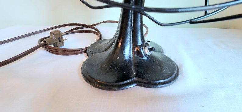 May include: Close-up of a vintage black electric fan. The fan has a round base with a three-leaf design, a black cage, and a brown power cord. The on/off switch is visible on the base.