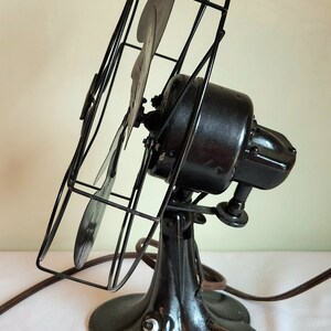 May include: An antique black electric fan with a cage and three blades. The fan has a decorative base and a brown power cord with a two-prong plug. The fan is angled slightly to the left.