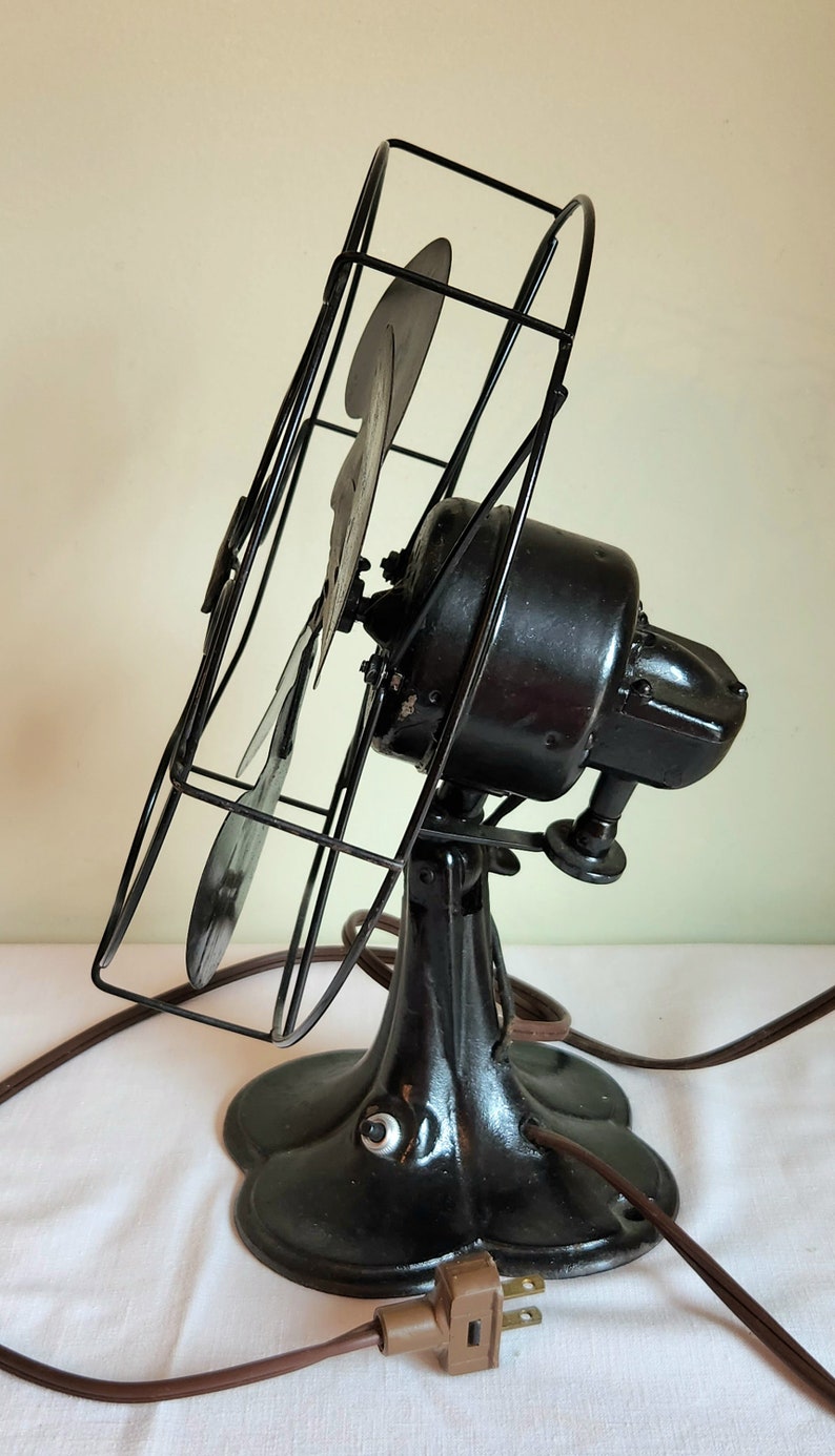 May include: An antique black electric fan with a cage and three blades. The fan has a decorative base and a brown power cord with a two-prong plug. The fan is angled slightly to the left.