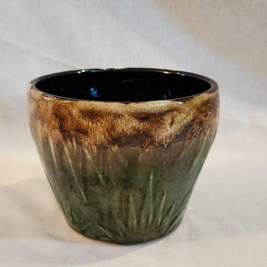 May include: A handmade ceramic bowl with a dark brown interior and a textured exterior. The bowl's rim is dark brown, transitioning to a lighter brown and green base. The green base has a grass-like texture.