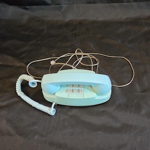May include: A light blue landline telephone with a matching coiled handset cord and a separate thin gray telephone line cable. The base features a rectangular keypad with white buttons, including standard numbers and symbols for dialing.