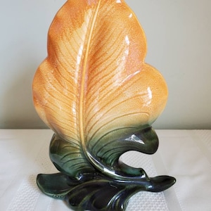 May include: A ceramic leaf-shaped vase with a yellow and green glaze. The vase is in the shape of a single leaf with a curled stem and a base that resembles a leaf.
