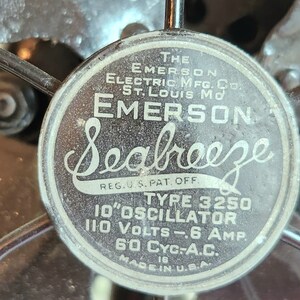 May include: Close-up of a vintage Emerson Seabreeze fan. The fan's central emblem displays the brand name and technical specifications, including "10" OSCILLATOR" and "110 VOLTS". The fan is made in the U.S.A.