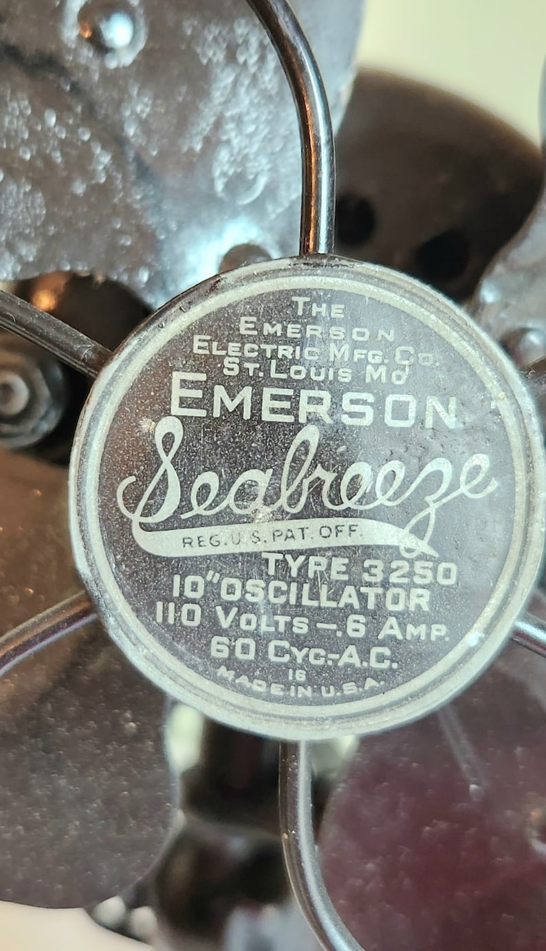 May include: Close-up of a vintage Emerson Seabreeze fan. The fan's central emblem displays the brand name and technical specifications, including "10" OSCILLATOR" and "110 VOLTS". The fan is made in the U.S.A.