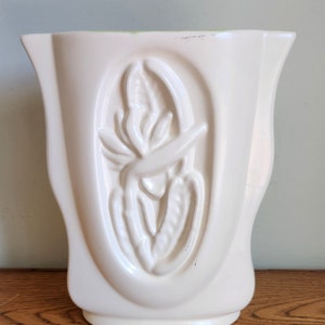 May include: A white ceramic vase with an embossed design of a tropical flower. The vase has a rounded, oval shape and a wide opening.