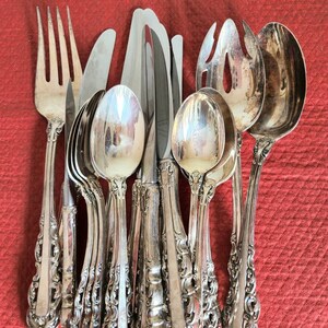 May include: An assortment of antique silverware, including forks, knives, and spoons, arranged on a red surface. The silverware has ornate, detailed handles and a shiny, reflective surface. The collection includes serving spoons and forks.